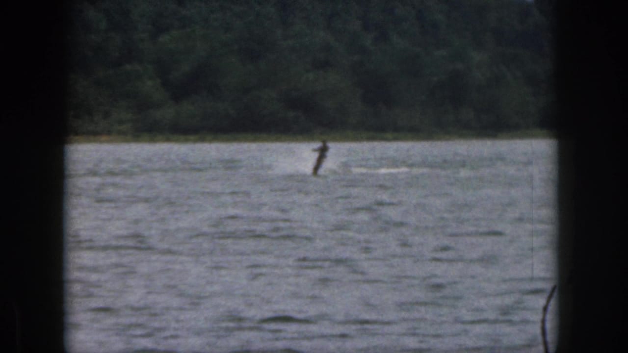 1961 Michigan 8mm frame showing a distant water skier on a lake, seen through dark foreground edges with trees beyond.