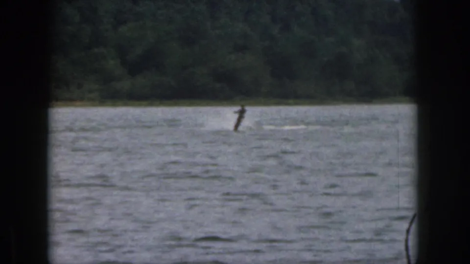 1961 Michigan 8mm frame showing a distant water skier on a lake, seen through dark foreground edges with trees beyond.