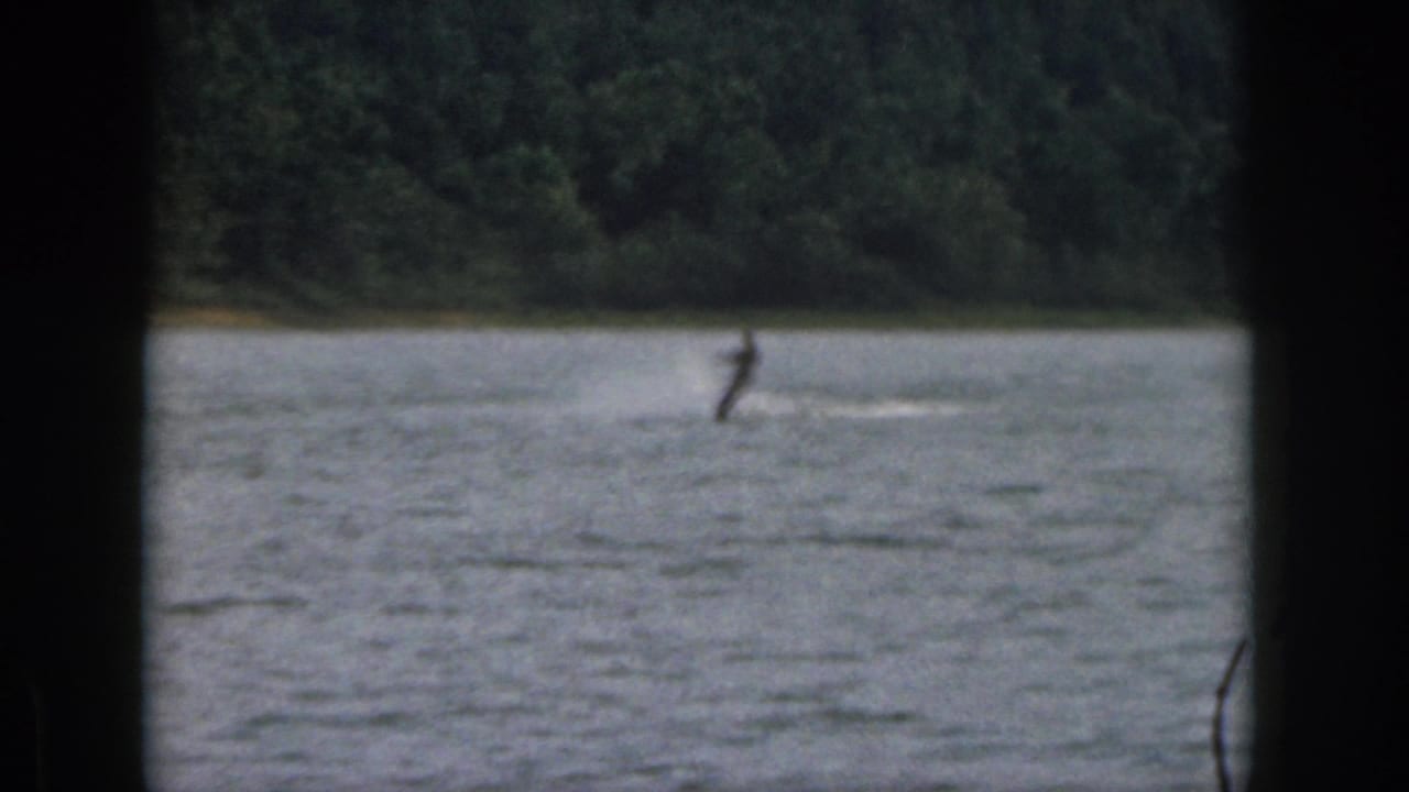 A distant skier passes across a Michigan lake in dim 8mm footage, with a dark foreground frame and forested shore visible.