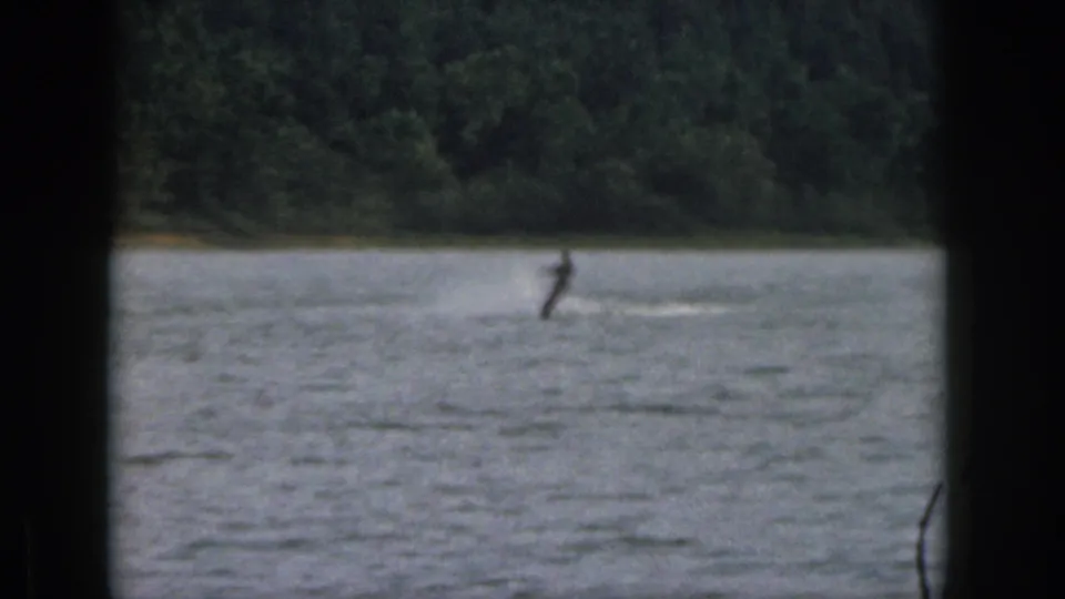 A distant skier passes across a Michigan lake in dim 8mm footage, with a dark foreground frame and forested shore visible.