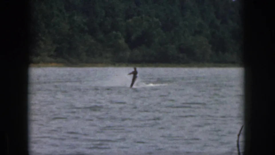 1961 home-movie still of a lone water skier on a wooded Michigan lake, partly obscured by a dark foreground frame.