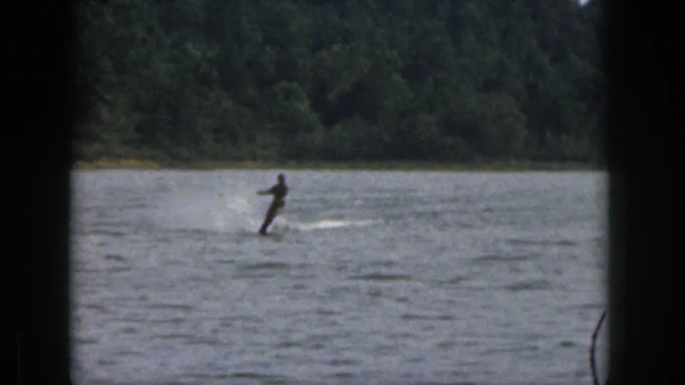 Distant 1961 Michigan lake view of a man water skiing, with spray behind him and a wooded shoreline in the background.