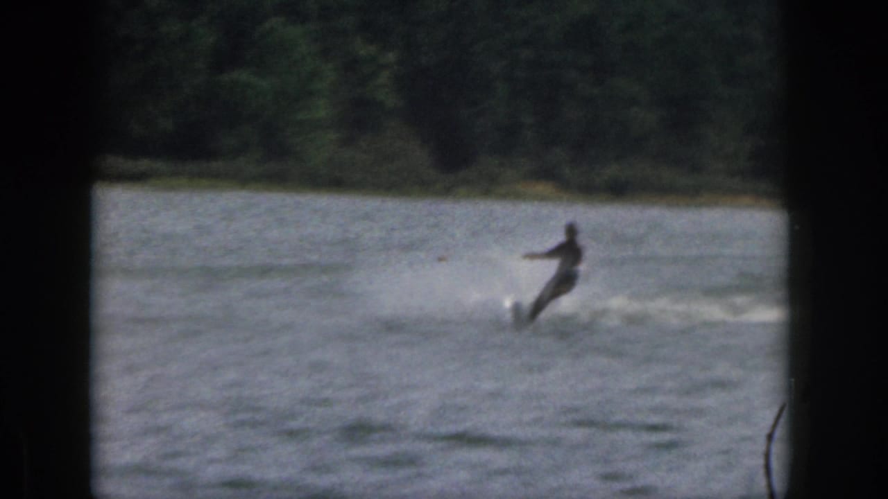 The skier leans into a turn on the lake, leaving a pale spray trail in the 1961 Michigan home-movie footage.