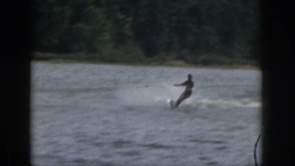The skier leans into a turn on the lake, leaving a pale spray trail in the 1961 Michigan home-movie footage.