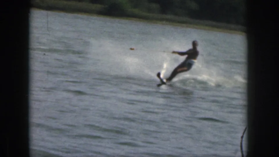 Closer 8mm view of the adult skier balancing on one ski as spray breaks beside him on a Michigan lake.