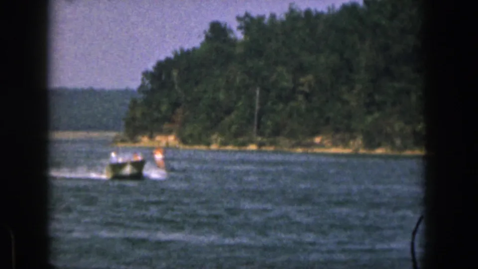 1961 Michigan lake recreation frame with a speedboat towing a water skier past a wooded shore in soft 8mm focus.