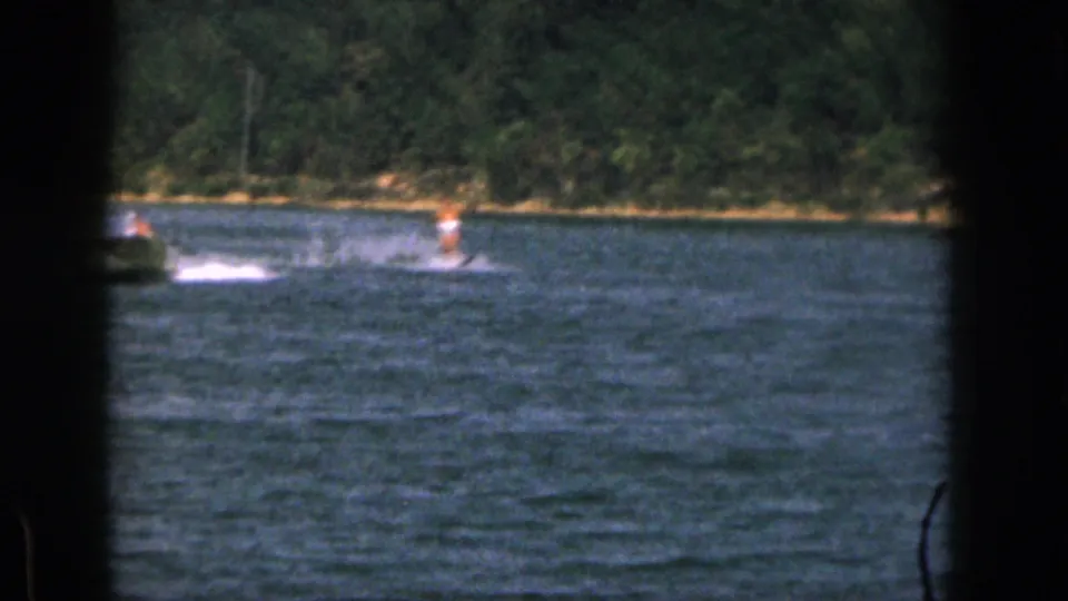 Distant water skier in a bright orange top rides behind a partly cropped boat on a Michigan lake in 1961 home-movie footage.