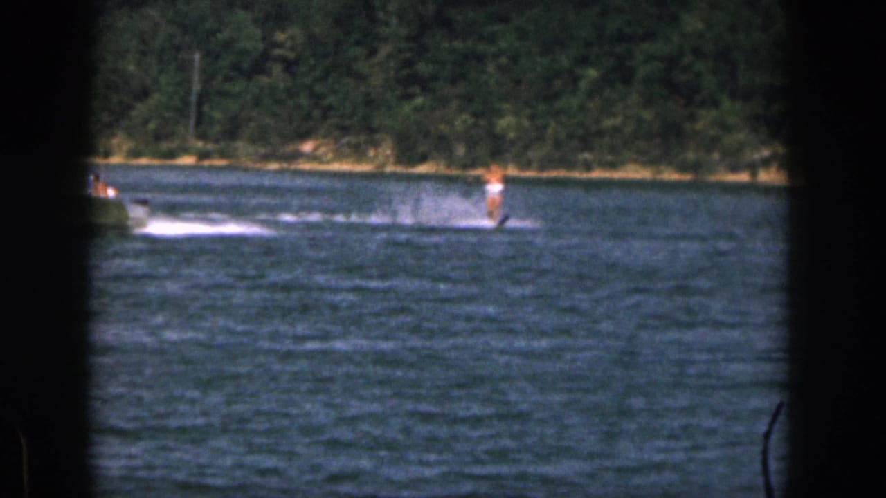 1961 Michigan home-movie frame showing a distant man water skiing behind a small boat on a river bordered by trees.