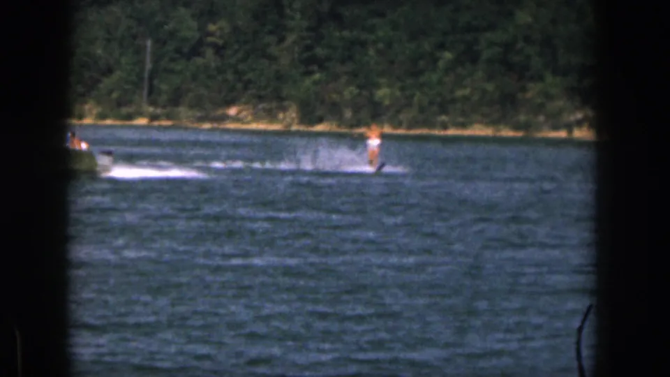 1961 Michigan home-movie frame showing a distant man water skiing behind a small boat on a river bordered by trees.
