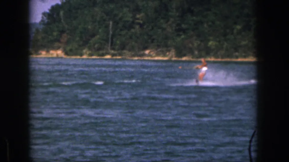 Closer 1961 Michigan home-movie view of the skier leaning on the water with forested shoreline in the background.