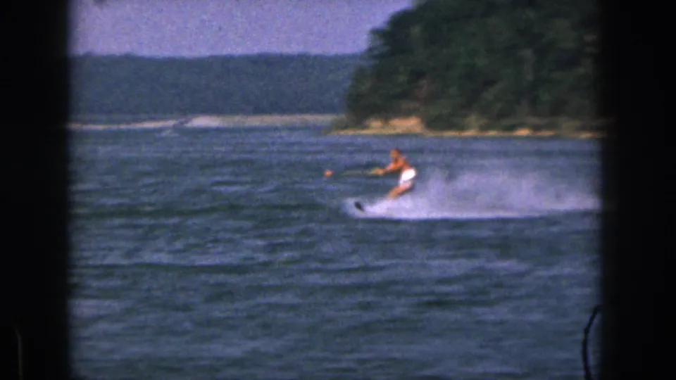 1961 Michigan frame showing the skier low over the water near a wooded bend, with spray trailing across the river surface.