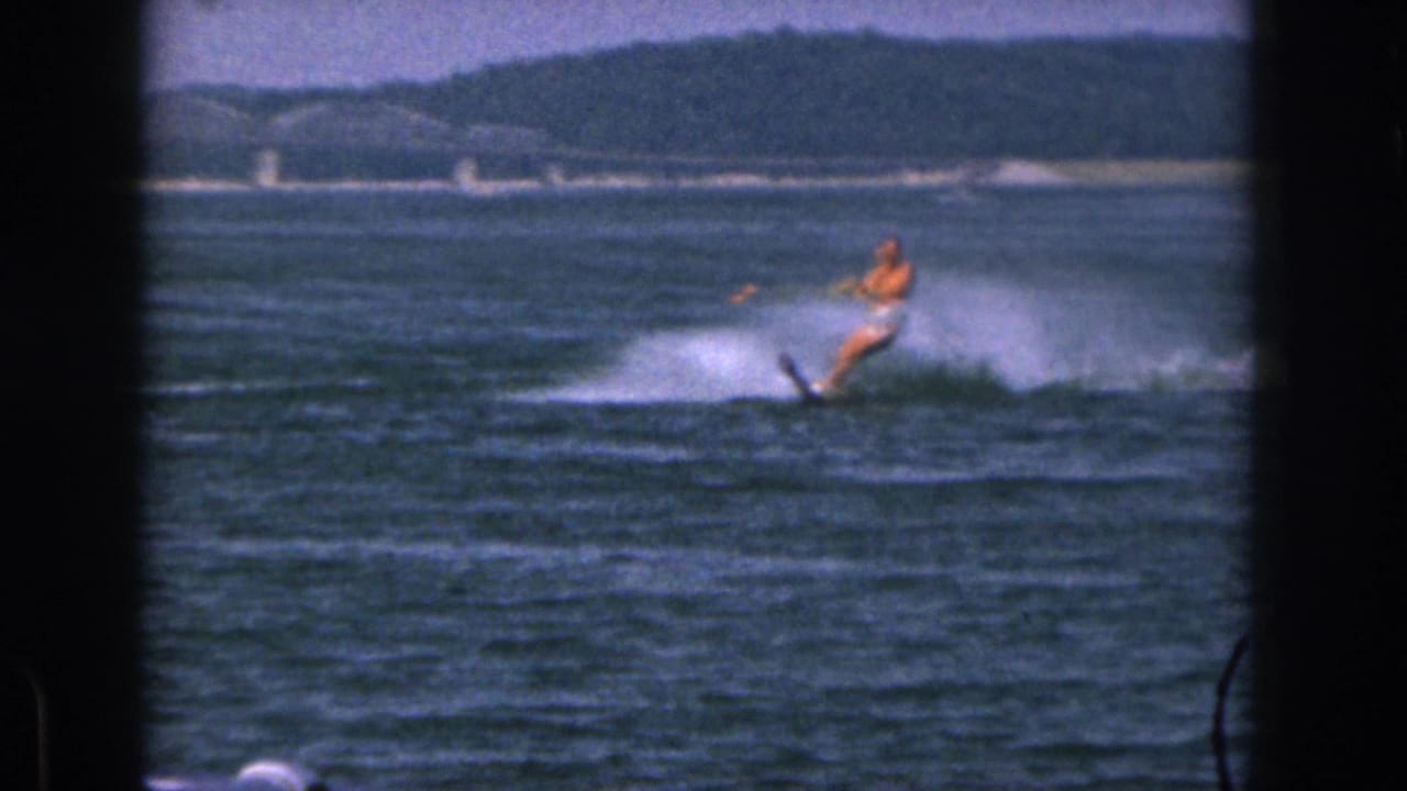 In 1961 Michigan home-movie footage, a man water skis across a lake at a distance with white spray, shoreline, and a bridge-like structure behind him.