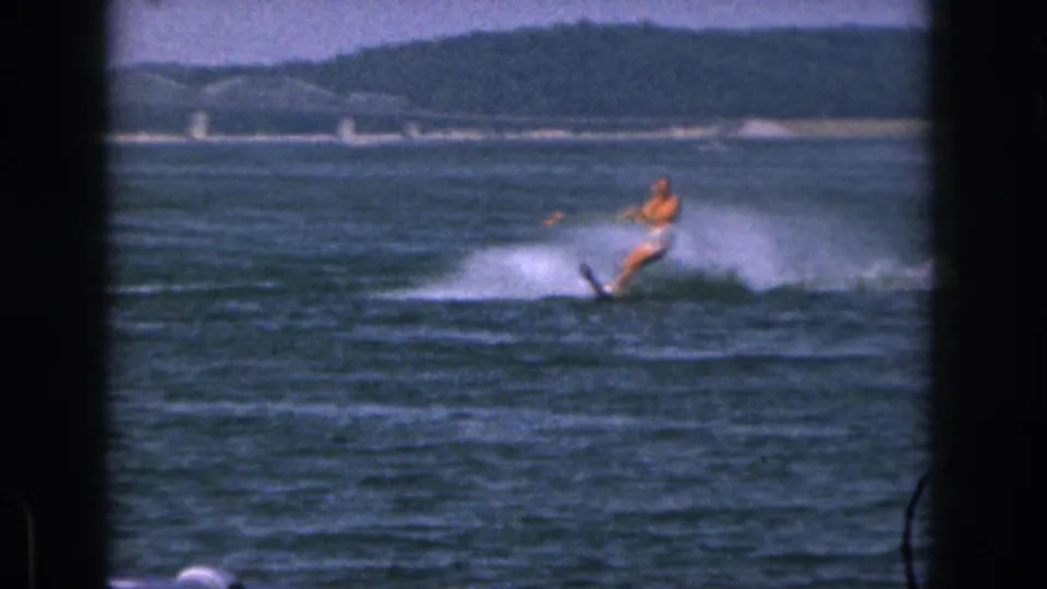 In 1961 Michigan home-movie footage, a man water skis across a lake at a distance with white spray, shoreline, and a bridge-like structure behind him.