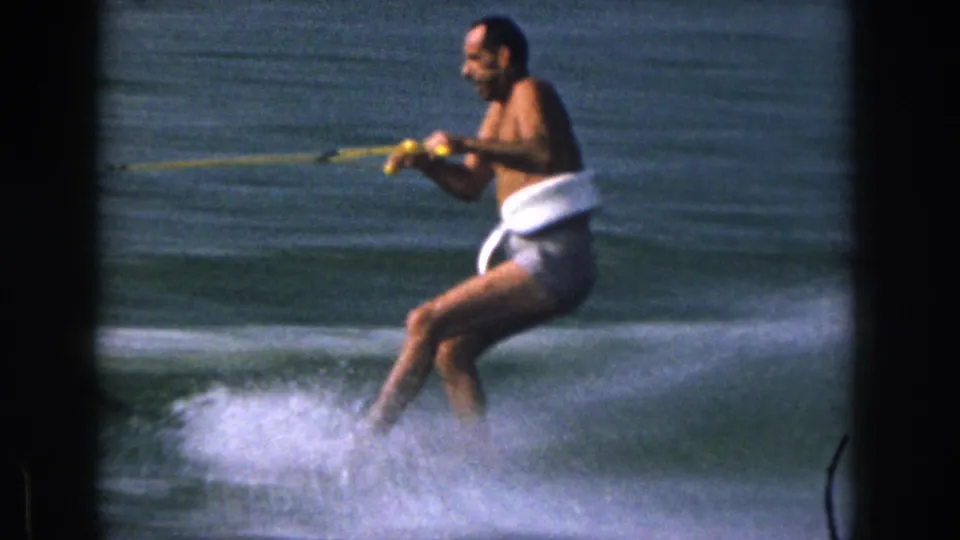 A closer side view shows the adult skier in white swim trunks leaning into the tow rope on a 1961 Michigan lake.