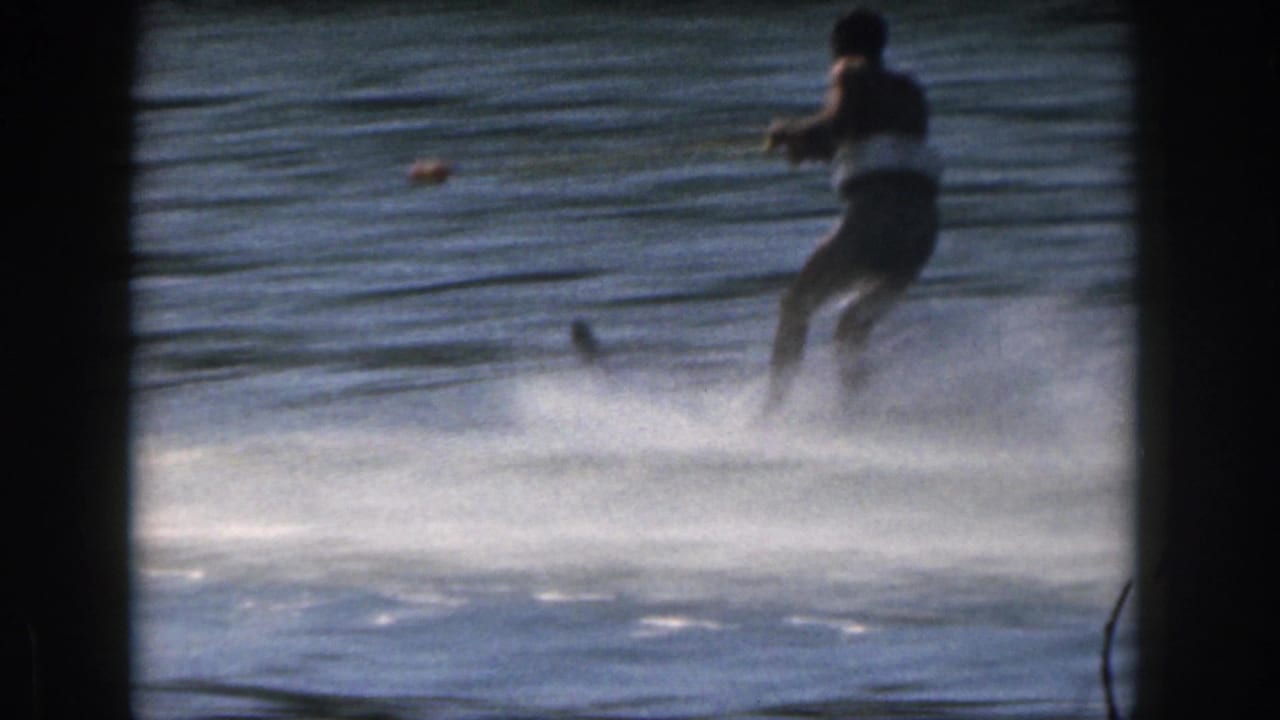 In 1961 Michigan 8mm footage, a man water skis on a lake while holding a tow rope, with white spray and projector borders visible.