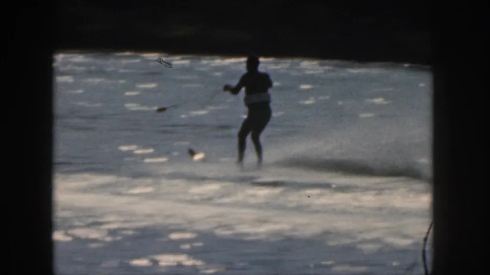 A distant man water skis across a Michigan lake in 1961 home-movie footage, with sunlight on the water and heavy film-frame borders.