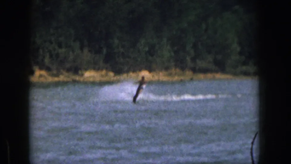 A distant water skier crosses a Michigan lake in 1961, with white wake and a tree-lined shore visible beyond the water.