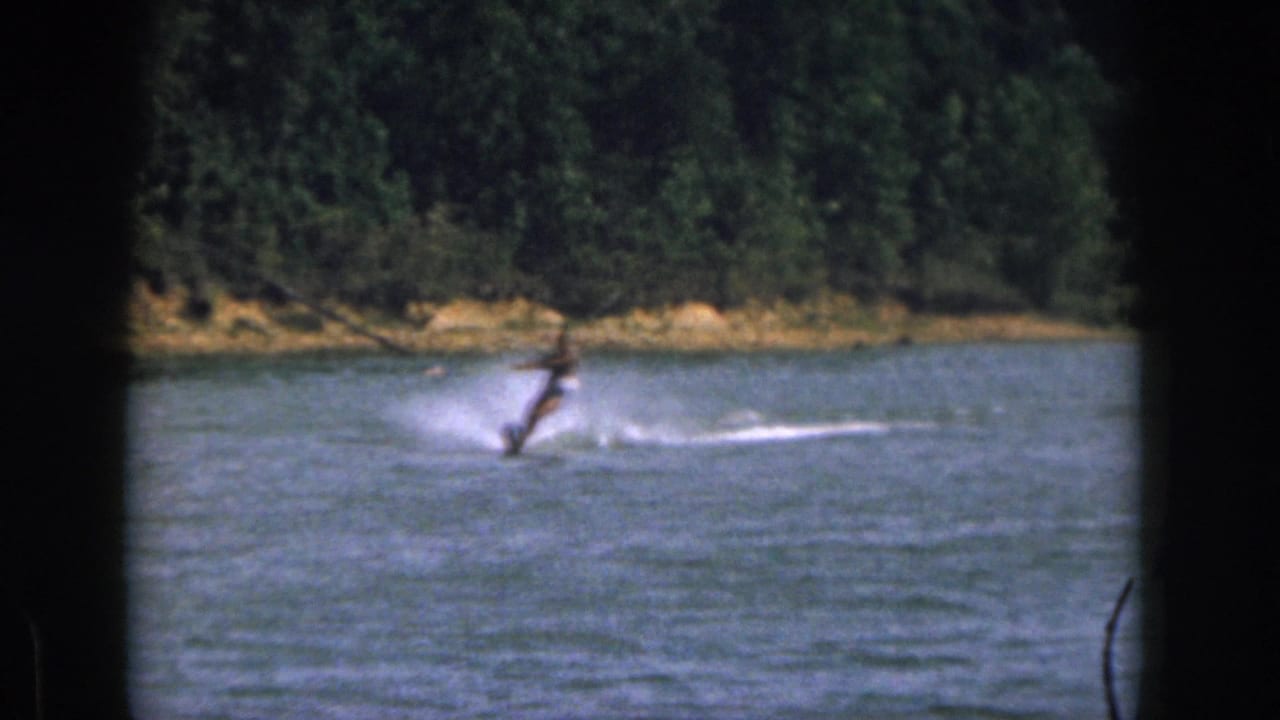 1961 Michigan home-movie frame of a man on a single water ski moving across calm lake water near a tree-lined shore.