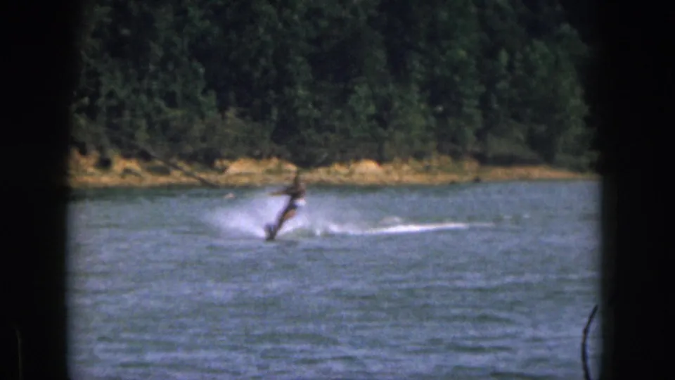 1961 Michigan home-movie frame of a man on a single water ski moving across calm lake water near a tree-lined shore.