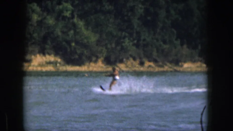 1961 Michigan frame showing a distant man water skiing on a calm lake, framed by dark film borders and wooded shore.