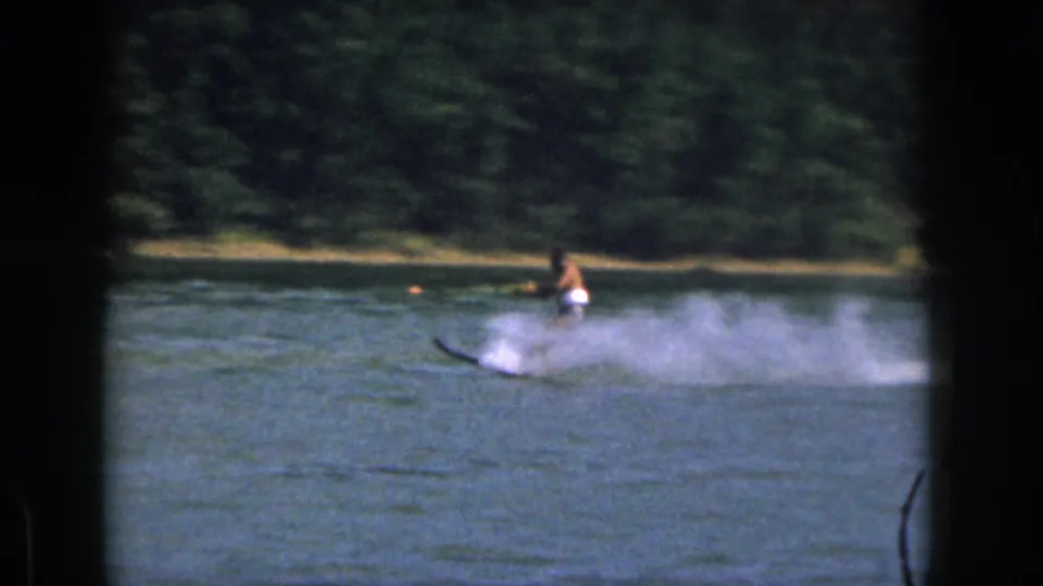 1961 Michigan home-movie still of a man water skiing through a larger spray cloud on an inland lake.