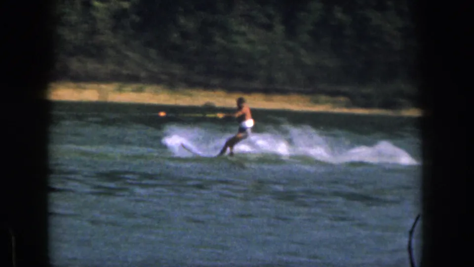 1961 Michigan frame of a man water skiing low across the water with wooded shoreline and sandy bank in the background.