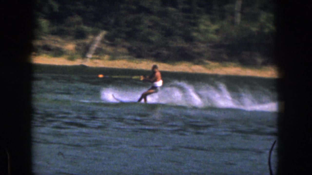 1961 Michigan 8mm frame showing a man in white swim trunks water skiing across a lake near a wooded shoreline.