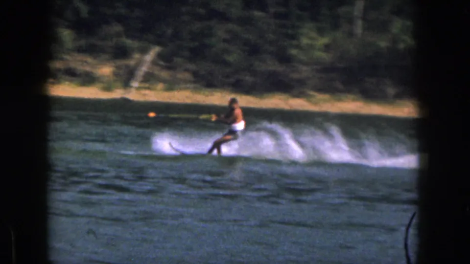 1961 Michigan 8mm frame showing a man in white swim trunks water skiing across a lake near a wooded shoreline.