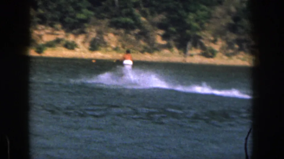 Distant 1961 Michigan 8mm view of a water skier moving away from camera across a lake with wooded shore behind.