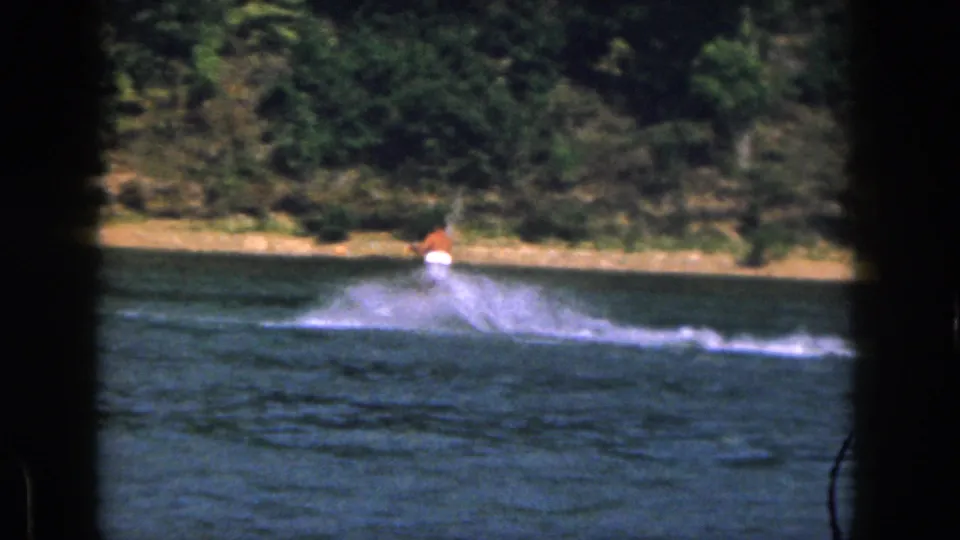 1961 Michigan home-movie frame showing a distant man water skiing along a wooded lakeshore with a long spray wake.