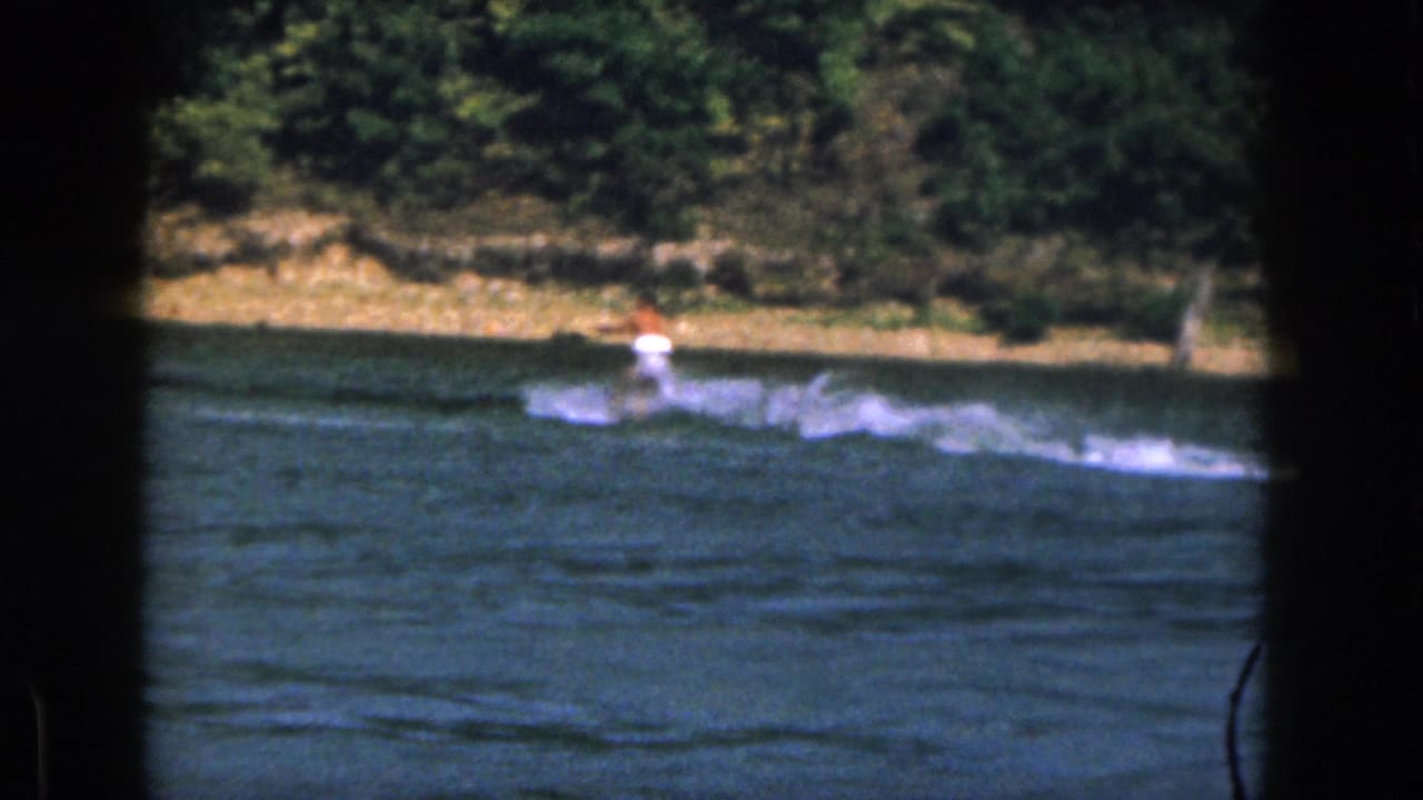1961 Michigan 8mm frame showing a distant water skier moving across a lake with spray behind him and trees along the shore.