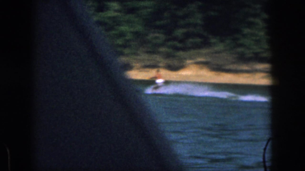 1961 Michigan home-movie frame with a small water-sport rider crossing the lake, partly obscured by dark projected-frame edges.