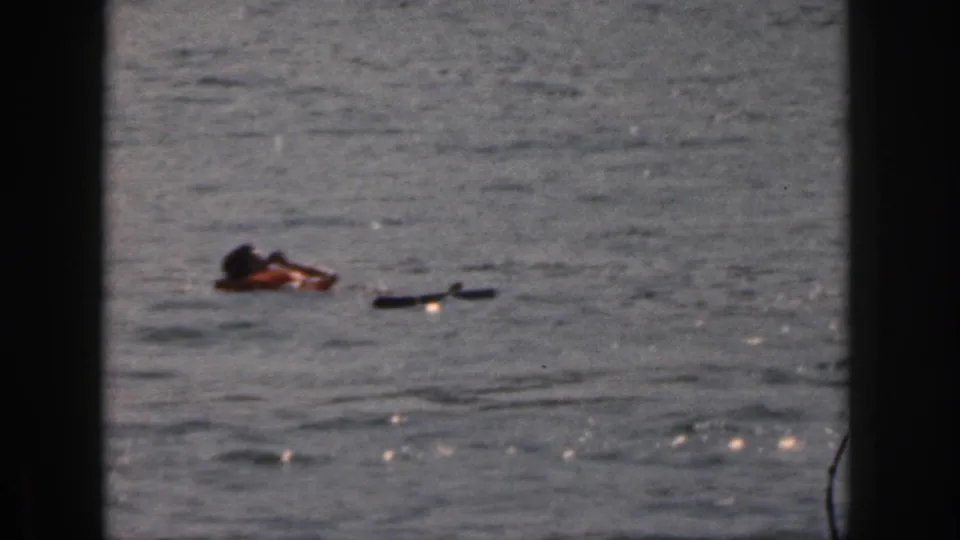 1961 Michigan home-movie frame of an adult swimmer in a life jacket moving through lake water beside a dark floating object.