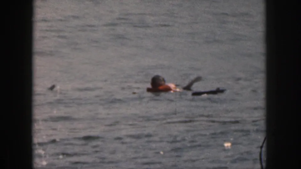 1961 Michigan 8mm still showing an adult partly submerged while swimming near a dark paddle-like object in open water.
