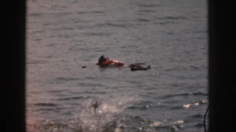 1961 Michigan 8mm frame showing a distant person in a life vest floating on rippled water, with splash marks near the foreground.
