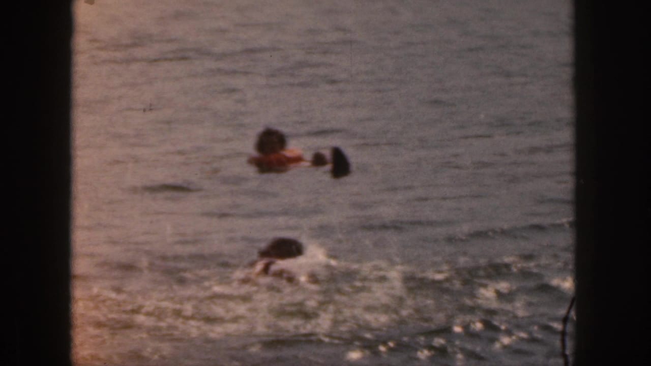 1961 Michigan 8mm home-movie frame showing two distant swimmers in open lake water with dark frame edges visible.
