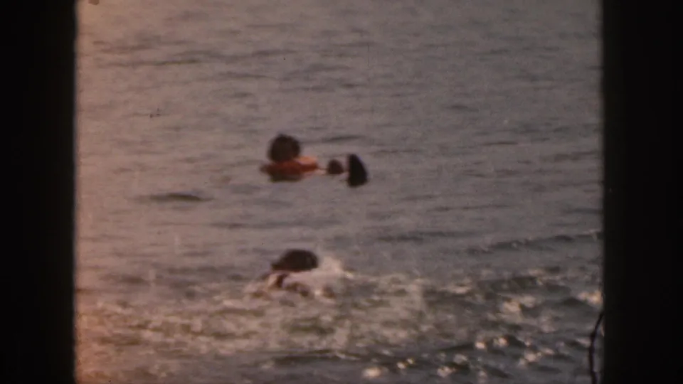 1961 Michigan 8mm home-movie frame showing two distant swimmers in open lake water with dark frame edges visible.