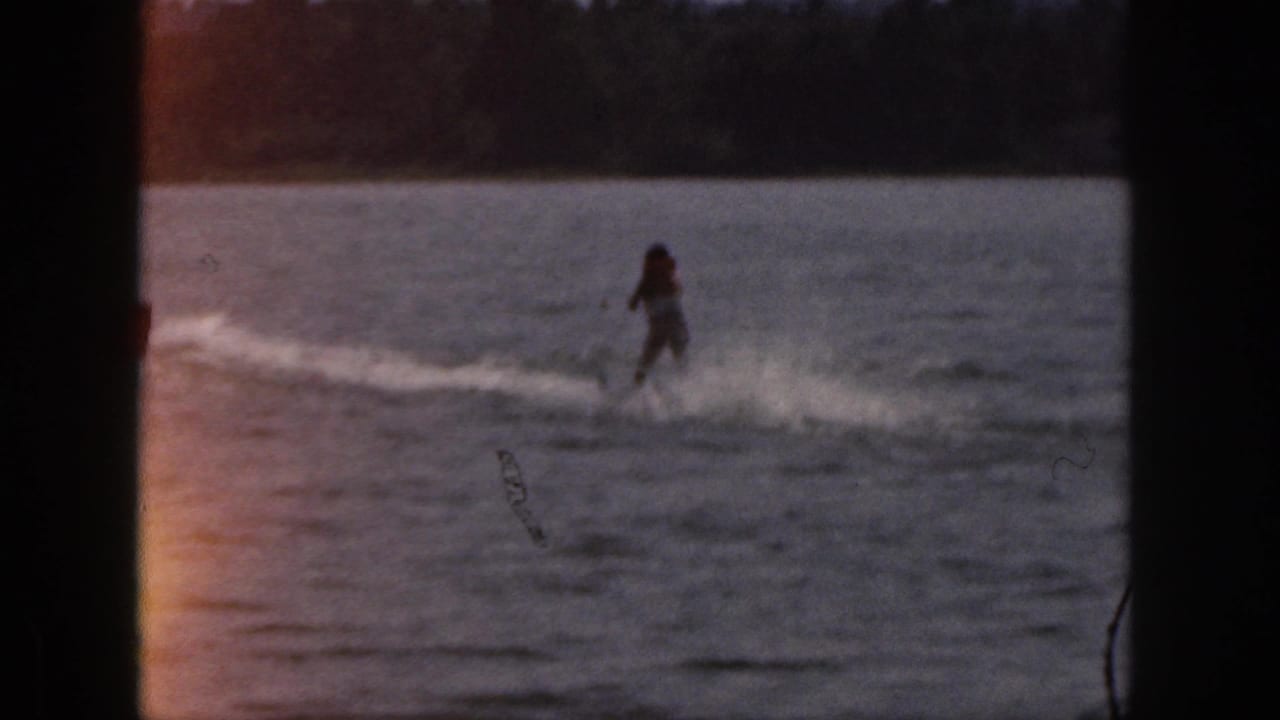 1961 Michigan home-movie frame showing a distant water skier crossing calm lake water in warm, low light.
