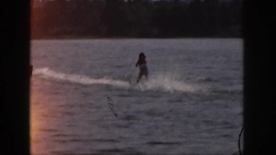 1961 Michigan home-movie frame showing a distant water skier crossing calm lake water in warm, low light.
