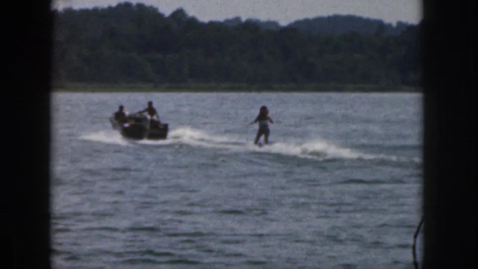 1961 Michigan lake scene with a small motorboat towing a distant water skier past a tree-lined shore.