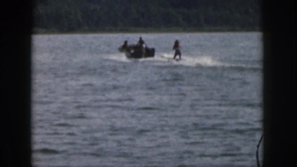 Distant 1961 Michigan home-movie view of a water skier following a small motorboat across rippled lake water.