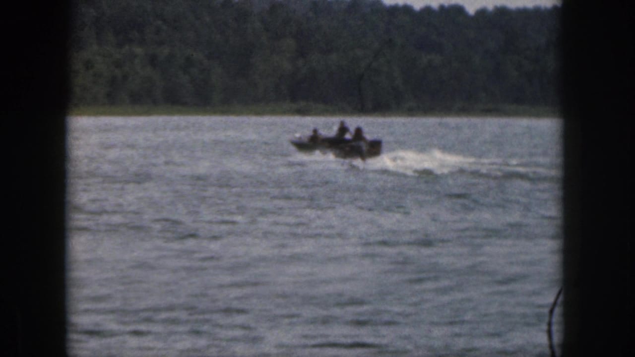 1961 Michigan home-movie frame of a boat carrying people across a lake, with wake and shoreline visible.