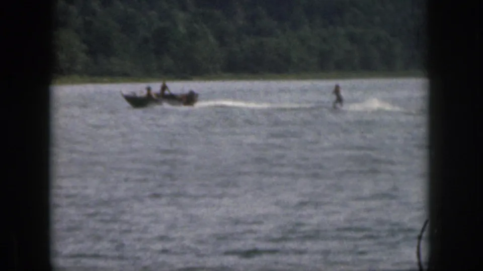 1961 Michigan home-movie frame with a boat towing a water skier across open lake water near trees.