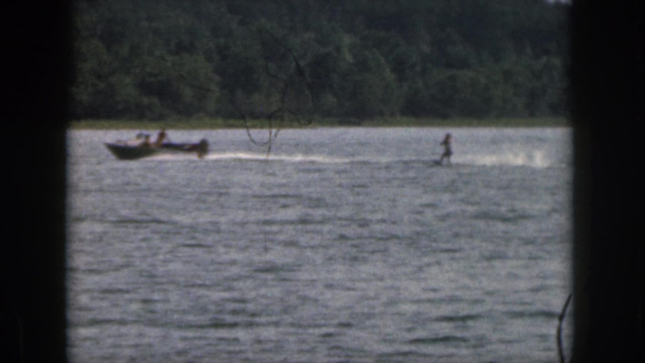 1961 Michigan home-movie frame showing a distant water skier pulled across a lake near a tree-lined shore.