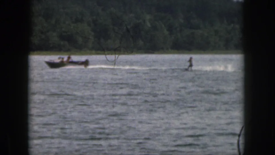 1961 Michigan home-movie frame showing a distant water skier pulled across a lake near a tree-lined shore.