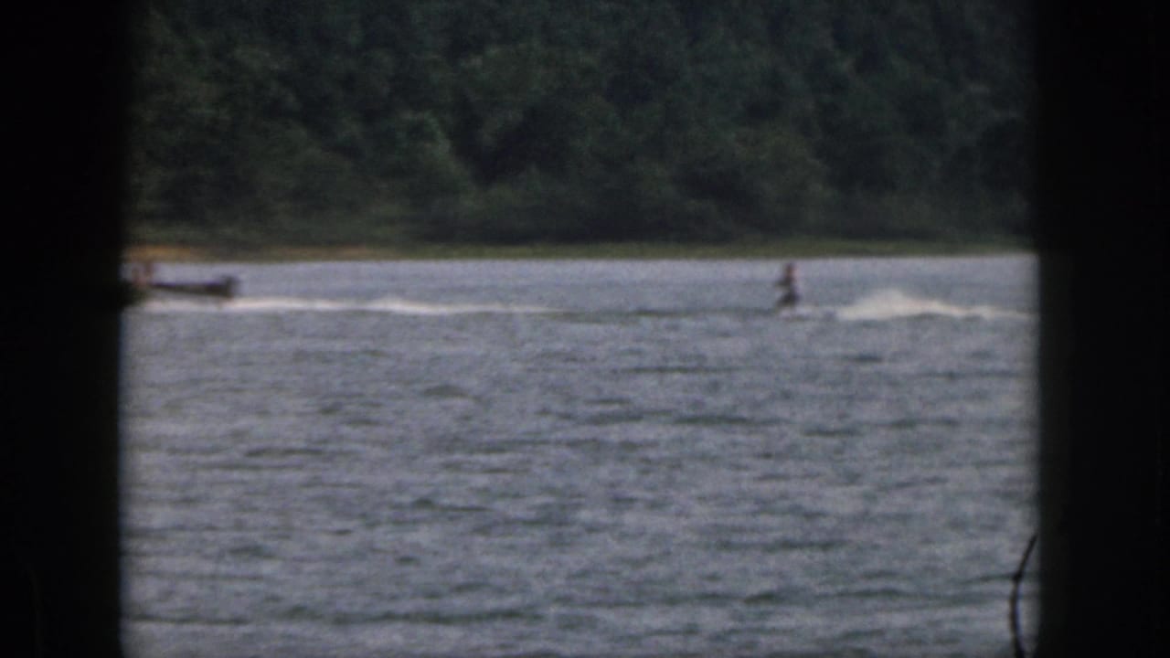Distant 1961 Michigan lake scene with a water skier being towed past a wooded shoreline.