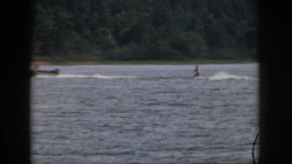 Distant 1961 Michigan lake scene with a water skier being towed past a wooded shoreline.