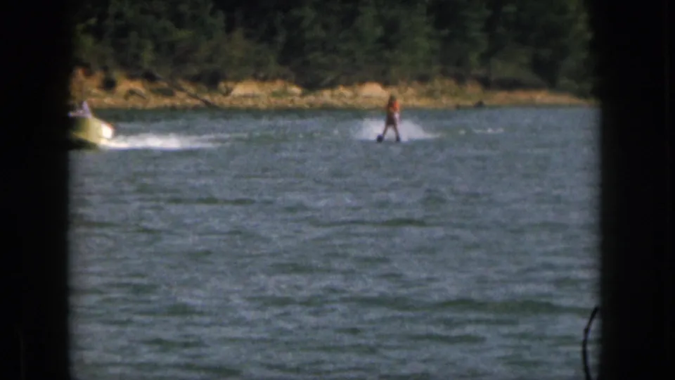 1961 Michigan home-movie frame showing a skier behind a partly visible motorboat near a wooded shore.
