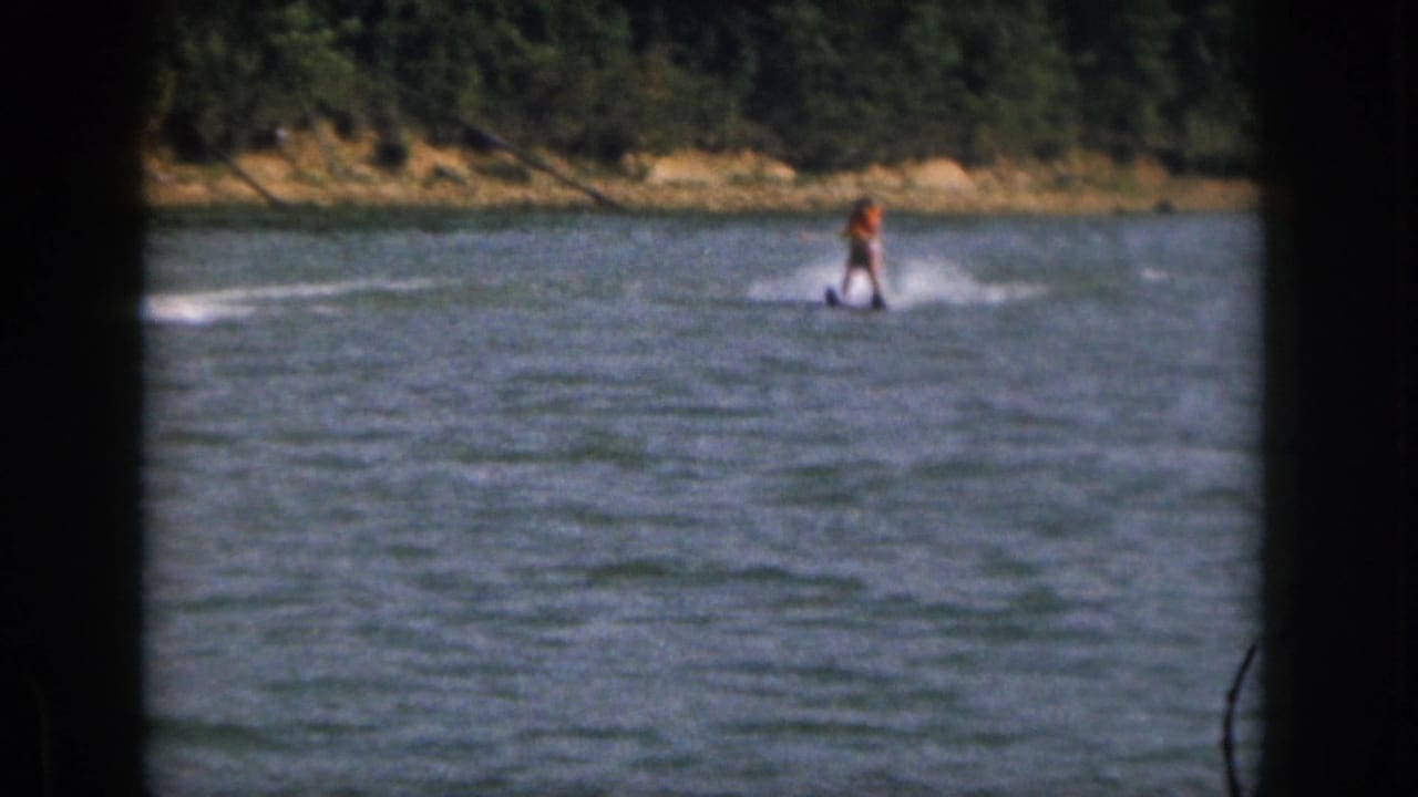 Water skier continues across open water with spray trailing behind and trees on shore.