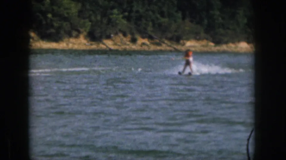 1961 home-movie frame of a distant water skier on a Michigan lake, seen through dark foreground framing with trees beyond the shore.